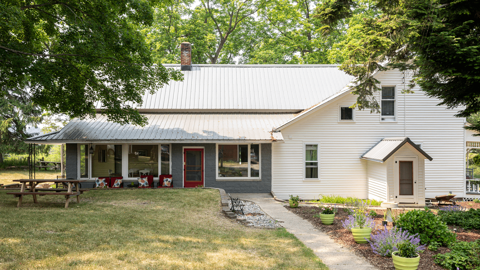 A white farmhouse with a metal roof and landscaped garden under green trees.
