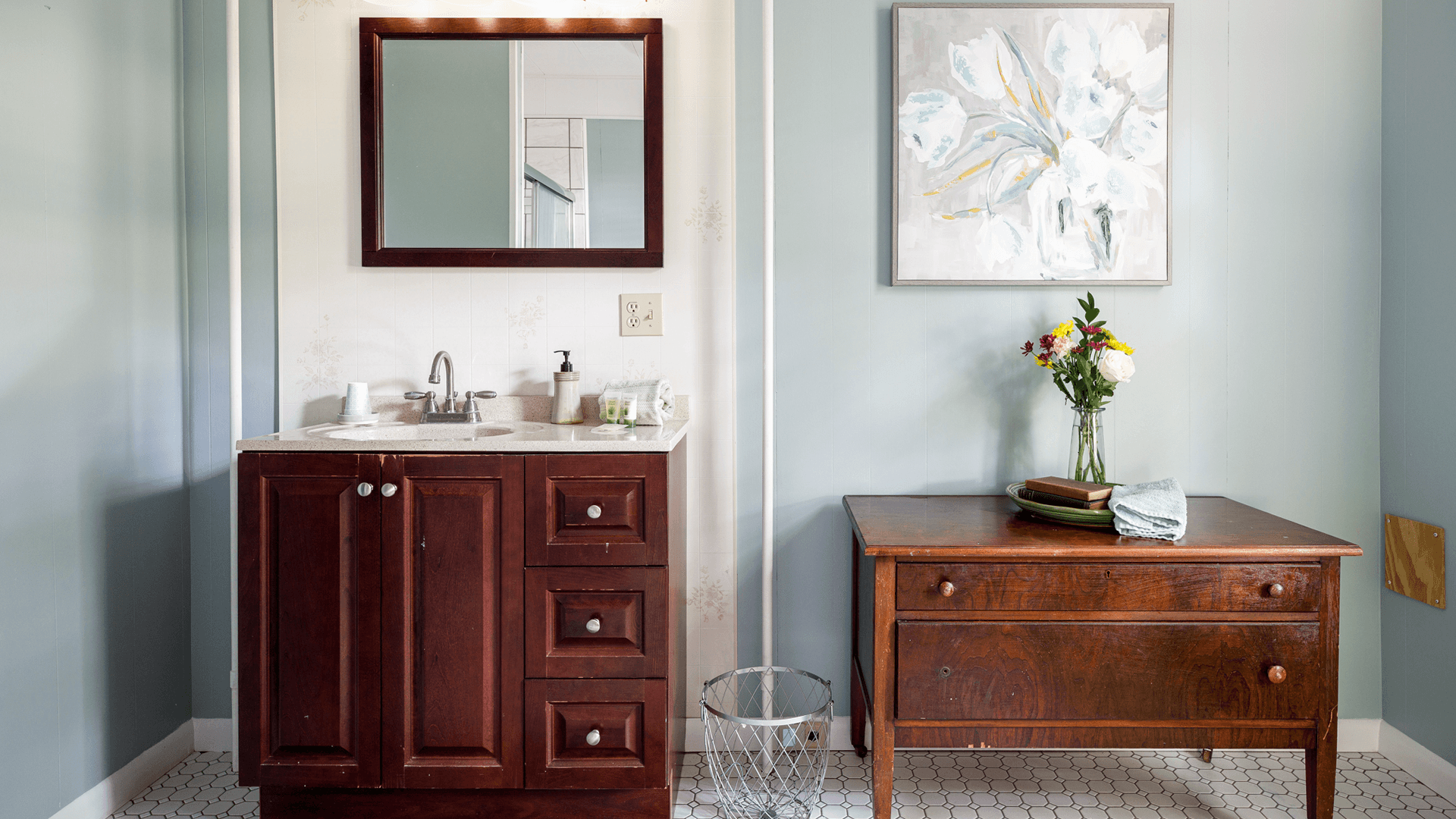 A modern bathroom featuring a wooden vanity, a round mirror, and a rustic wooden table with flowers.