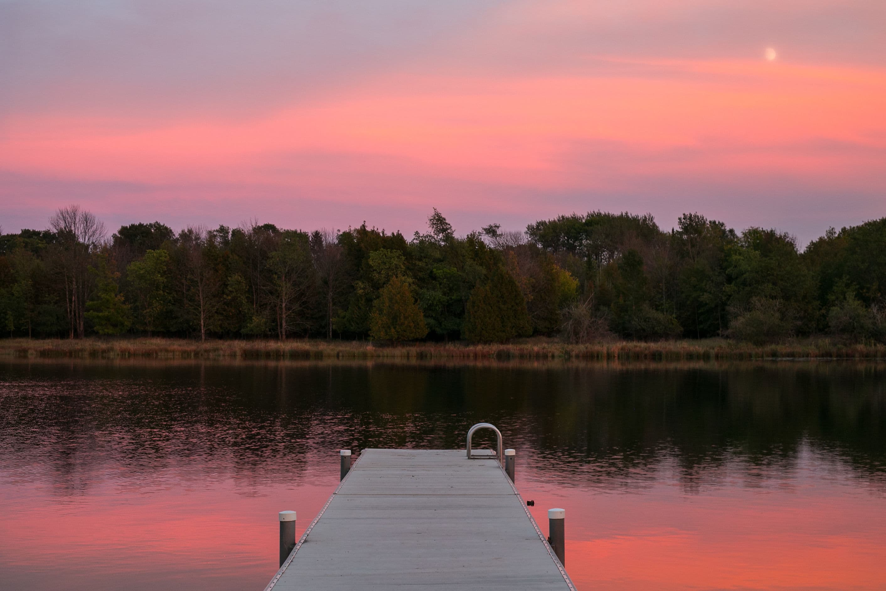 A wooden dock extends into a calm lake reflecting a pink and purple sky at sunset.