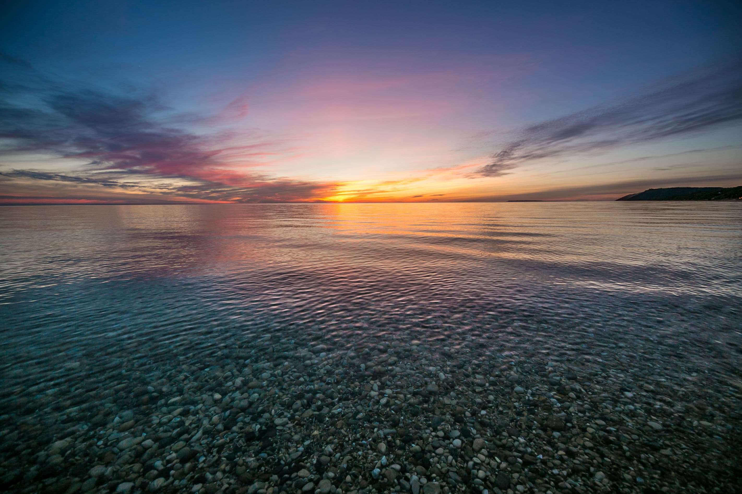 A tranquil sunset over a calm sea with visible pebbles in the foreground.