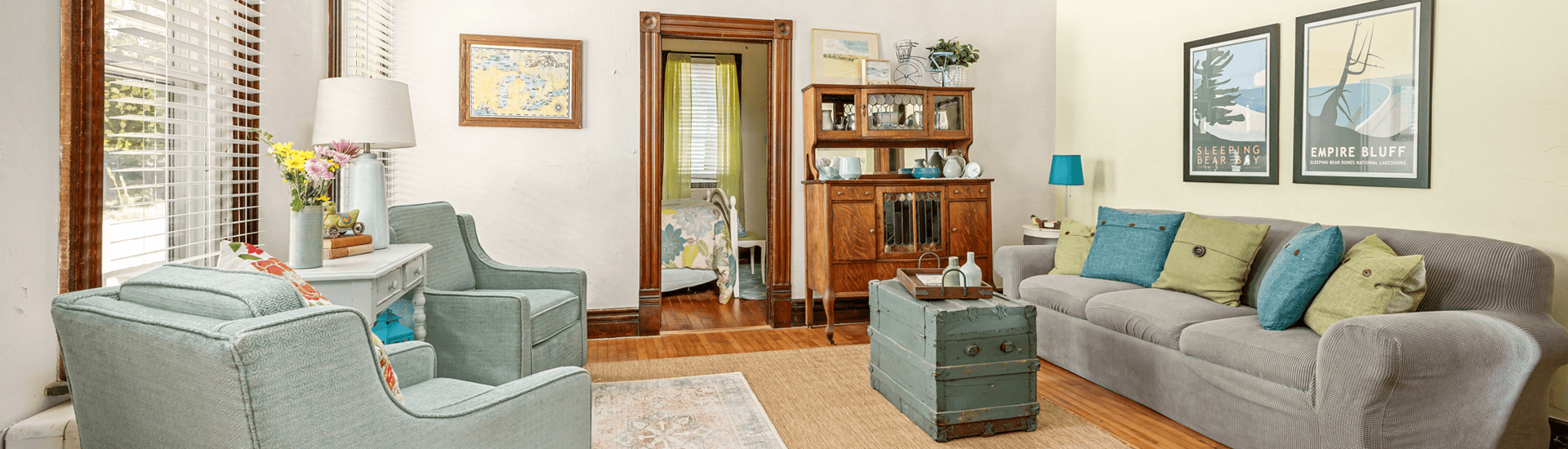 A cozy living room featuring a gray sofa, teal chairs, and a wooden cabinet, with colorful artwork on the walls.